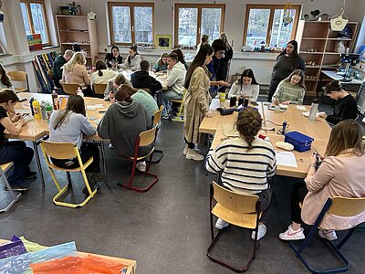 Eine Gruppe von Schülerinnen sitzt an mehreren Tischen in einem Klassenzimmer und arbeitet an kreativen Projekten mit verschiedenen Bastelmaterialien. Die Schülerinnen sind konzentriert bei der Arbeit, während andere durch den Raum gehen und Unterstützung anbieten. Der Raum ist gut beleuchtet und hat große Fenster.