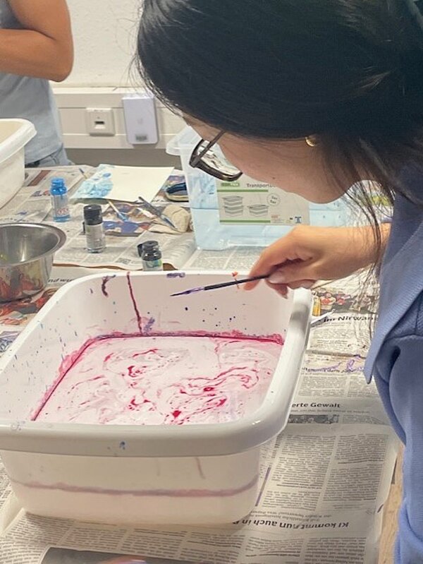 Eine Person mit Brille, schwarzem Haar und blauer Kleidung arbeitet konzentriert an einem Tisch. Sie hält einen Pinsel und bearbeitet eine weiße Kunststoffwanne, die mit roter und weißer Farbe gefüllt ist. Im Hintergrund sind weitere Materialien und Zeitungen als Unterlage zu sehen.