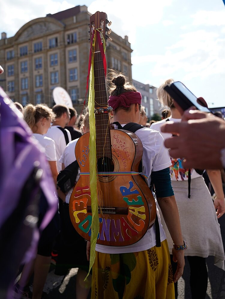 REWE Team Challenge 2024 Eine Frau mit langen, zu einem Dutt hochgebundenen Haaren, die von hinten in einer Menschenmenge gezeigt wird. Sie hat bunte bemalte Gitarre auf dem Rücken mit der Aufschrift "Heldenmacher - Erzieher, Sozialassistenten"