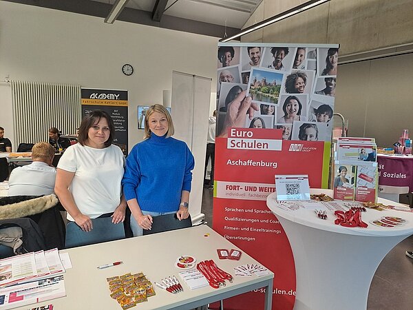 Auf der Weiterbildungsmesse Main.Fortschritt Frau Beller und Frau May strahlen am Messestand neben dem Roll-up der Euro-Schulen um die Wette in die Kamera. Auch einige Messegeschenke sind auf den Tischen zu sehen.