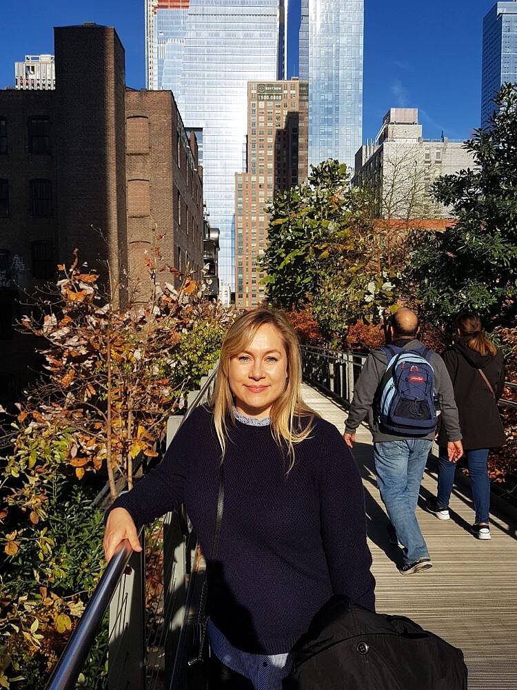 Alumni im Gespräch: Interview mit Cathrin Weinel Eine blonde Frau mit einem dunkelblauen Pullover steht auf der High Line in New York City und lehnt sich an das Geländer. Hinter ihr sind herbstlich gefärbte Bäume, Fußgänger sowie moderne Wolkenkratzer und ältere Backsteingebäude zu sehen. Die Sonne scheint, und der Himmel ist blau.