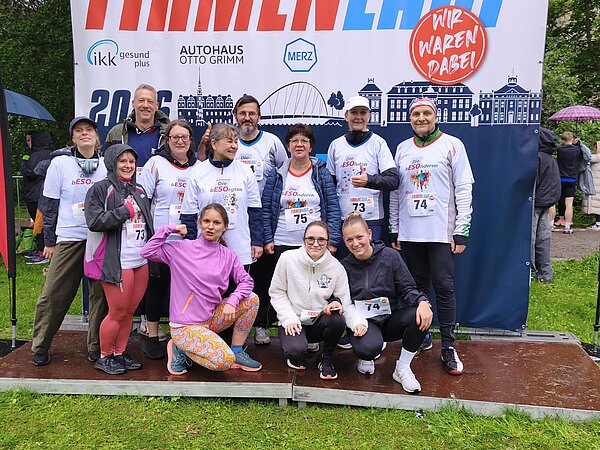 Gruppenfoto von Läufer*innen beim Firmenlauf vor einer Veranstaltungswand; die Teilnehmenden tragen Laufshirts mit Startnummern und posieren gemeinsam im Freien.