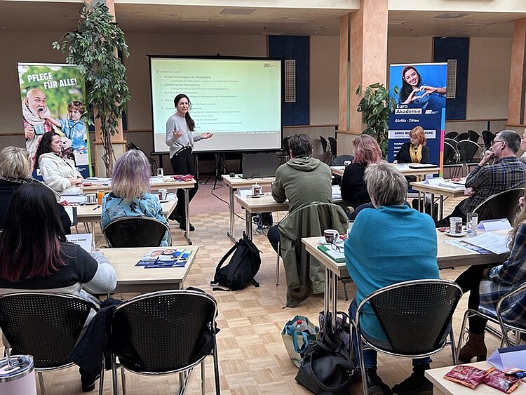 Großer bestuhlter Saal mit Tischen und Teilnehmern und Kooperationspartnern, die einem Vortrag zuhören, im Hintergrund eine Präsentation.