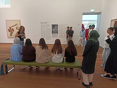 Gruppenbesuch in einem Museum: Eine Frau erklärt einer sitzenden und stehenden Gruppe von jungen Erwachsenen ein Kunstwerk an der Wand.