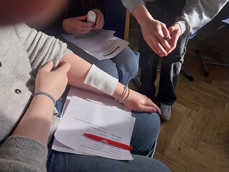Ein Arm mit einem weißen Verband liegt auf einem Blatt Papier, das auf einem Oberschenkel ruht. Ein Schüler hält einen Stift, eine andere Person reicht einen kleinen Gegenstand. Die Szene spielt in einem Innenraum mit Holzboden. Sonnenlicht fällt auf die Personen und die Unterlagen.