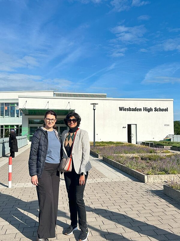 Zwei Personen stehen vor dem Eingang des modernen Gebäudes der Wiesbadener High School. Das Gebäude ist hell und von gepflegten Beeten mit Lavendel umgeben. Der Himmel ist klar und blau.
