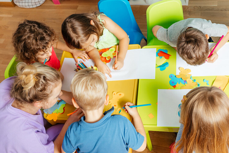 Berufsbegleitend Erzieher*in werden Eine Gruppe von Kindern sitzt an einem Tisch in einem Klassenzimmer und bastelt. Sie verwenden bunte Papierstücke und Buntstifte, während eine Erzieherin ihnen dabei hilft. Der Tisch ist mit verschiedenen Farben und Bastelmaterialien bedeckt.