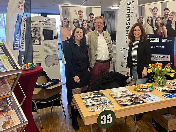 Messestand der Euro Akademie Hohenstein-Ernstthal auf der Ausbildungsmesse in der Stadthalle Limbach-Oberfrohna. Im Vordergrund ein rechteckiger Tisch mit Informationsmaterialien, Flyern und Broschüren, dekoriert mit einem Strauß gelber und roter Tulpen sowie kleinen Schalen mit Süßigkeiten. Auf dem Tisch liegt ein großes Schild mit der Zahl 35. Links ein Prospektständer mit mehreren Fächern, gefüllt mit Flyern der Euro Akademie. Im Hintergrund Roll-ups mit Logos und Informationen zur Euro Akademie und zur Berufsausbildung. Drei Mitarbeitende der Euro Akademie stehen hinter dem Tisch, tragen Businesskleidung, lächeln in die Kamera und wirken einladend. Die Szenerie ist hell beleuchtet, der Boden besteht aus Holzparkett. Die Atmosphäre vermittelt einen professionellen und freundlichen Eindruck eines Messeauftritts.