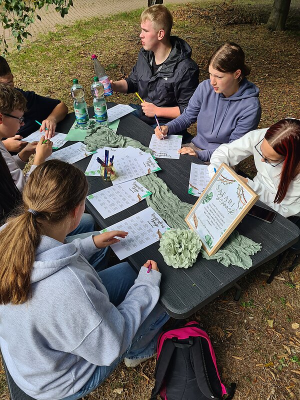 Kennenlerntag der Euro Akademie Rochlitz Mehrere Jugendliche sitzen an einem Tisch im Freien und spielen Kennenlern-Bingo. Auf dem Tisch liegen Blätter, Stifte, Wasserflaschen und ein dekorativer Schriftzug.