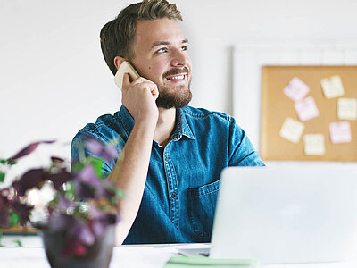 Das Bild zeigt eine Person in einem blauen Hemd, die am Telefon spricht. Sie sitzt an einem Schreibtisch mit einem weißen Laptop und grünen Papieren. Im Hintergrund ist ein unscharfes Notizbrett zu sehen.