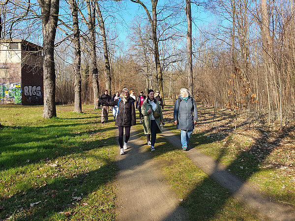 Exkursion in die Natur Die Gruppe spaziert entspannt auf einem Waldweg entlang. Im Vordergrund unterhalten sich drei Frauen, im Hintergrund sind weitere Teilnehmende zu sehen. Links steht ein altes Gebäude mit Graffiti.