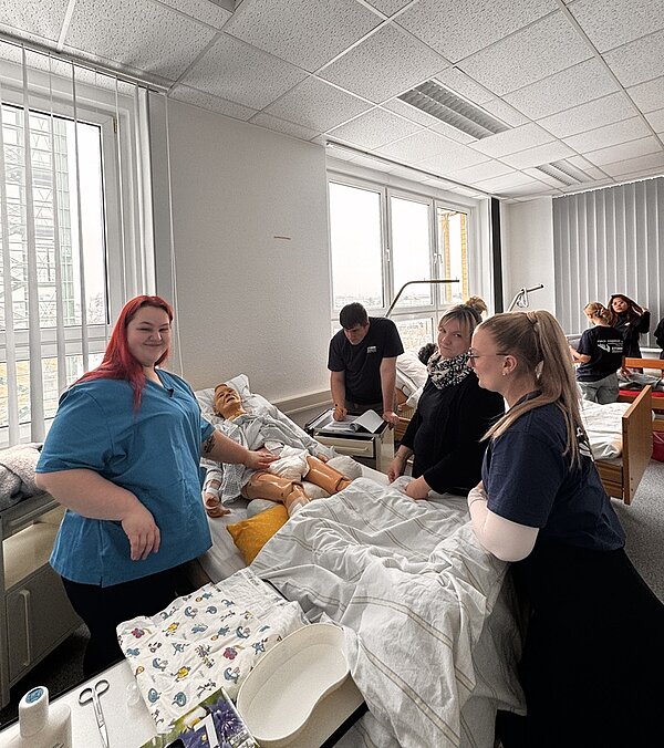 Mehrere Auszubildende und Lehrkräfte stehen um ein Krankenbett, auf dem eine lebensechte Übungspuppe liegt. Die Gruppe trägt dunkle Kleidung, eine Person trägt einen hellblauen Kittel. Auf dem Bett und dem Tisch liegen medizinische Utensilien wie Tücher und Schalen. Der Raum ist hell, mit großen Fenstern und weißen Wänden. Im Hintergrund sind weitere Betten und Übungspuppen zu sehen.