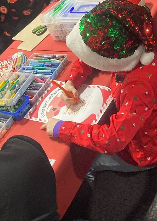 Ein Kind in einem roten Weihnachtspullover und einer Weihnachtsmütze sitzt an einem Tisch und malt ein Bild mit Buntstiften. Auf dem Tisch liegen verschiedene Malutensilien.