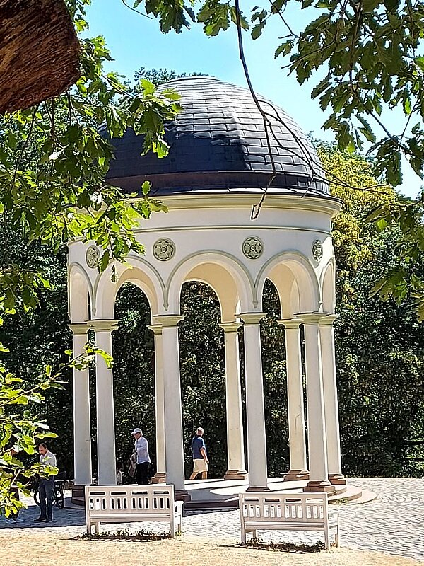 Ein runder, weißer Pavillon mit Säulen und Treppen, umgeben von üppigem Grün und Bäumen.