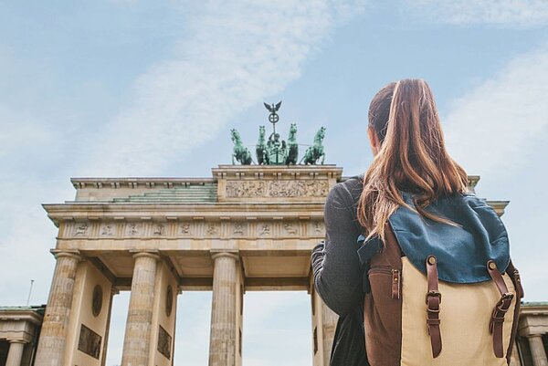 Eine Person mit langem, braunem Haar und einem blauen Rucksack mit braunen Lederakzenten steht mit dem Rücken zur Kamera. Die Person blickt auf das Brandenburger Tor in Berlin. Das Brandenburger Tor ist ein klassizistisches Bauwerk mit Säulen und einer Quadriga (ein von vier Pferden gezogener Wagen) auf der Spitze. Der Himmel ist leicht bewölkt und hell.