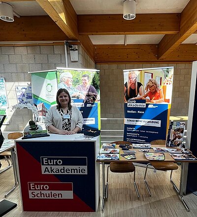 Mitarbeitende der Euro Akademie Meißen und Riesa steht gut gelaunt und lächelnd am Messestand, der mit Euro Akademie Roll-Up, Stühlen, Tischen, einem Stehtisch ausgestattet ist.
