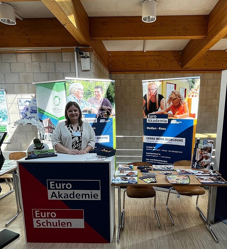 Erfolgreiche Teilnahme an der Messe „Radebeul jobbt“ Mitarbeitende der Euro Akademie Meißen und Riesa steht gut gelaunt und lächelnd am Messestand, der mit Euro Akademie Roll-Up, Stühlen, Tischen, einem Stehtisch ausgestattet ist.
