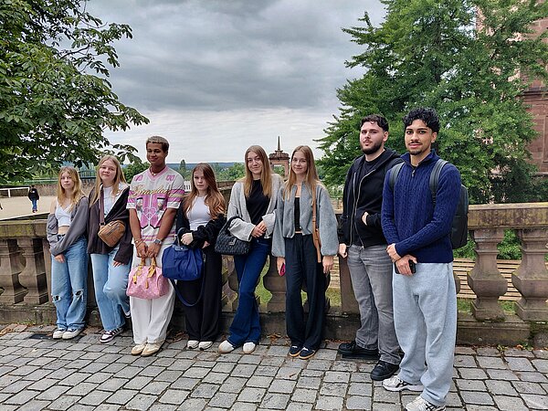 Eintauchen in die Geschichte Aschaffenburgs Eine Gruppe junger Menschen steht auf einem gepflasterten Platz vor einer Balustrade mit Blick auf eine historische Stadtlandschaft. Der Himmel ist bewölkt, im Hintergrund sind Bäume und ein Kirchturm zu sehen.