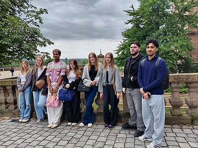 Eine Gruppe junger Menschen steht auf einem gepflasterten Platz vor einer Balustrade mit Blick auf eine historische Stadtlandschaft. Der Himmel ist bewölkt, im Hintergrund sind Bäume und ein Kirchturm zu sehen.