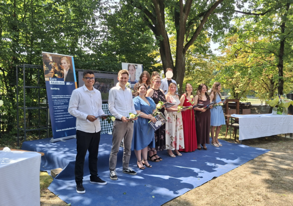 Gruppe von Absolventinnen und Absolventen steht auf einer Bühne im Freien und hält Rosen sowie Mappen mit Urkunden in den Händen. Die Veranstaltung findet unter großen Bäumen statt, im Hintergrund stehen Werbebanner der Euro Akademie Bamberg.