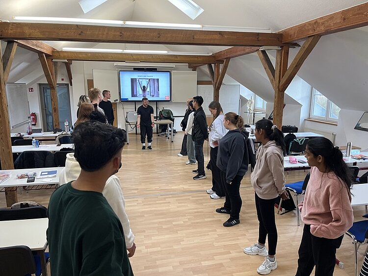 Auszubildende zur Pflegefachkraft der Euro Akademie Pößneck nehmen im Rahmen eines Gesundheitstages an einer praktischen Gruppenübung teil. Die Teilnehmenden hocken sich in zwei Reihen gegenüber auf den Boden und folgen den Anweisungen eines Trainers, der eine gesundheitsfördernde Übung demonstriert. Der lichtdurchflutete Seminarraum mit Holzstützen und Präsentationstechnik unterstreicht den praxisorientierten Charakter des Workshops. Die Szene vermittelt Teamarbeit, Bewegung, Gesundheitsprävention und Reflexion über den eigenen Umgang mit digitalen Medien.