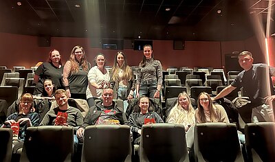 Ein dunkler Kinosaal mit Sesseln. In den Reihen stehen und sitzen 13 junge Menschen und blicken lächelnd in die Kamera.