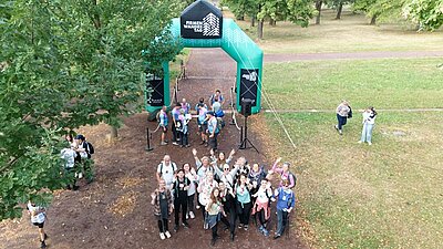Aus der Vogelperspektive fotografiertes Gruppenbild der Mitarbeitenden der Euro-Schulen und Euro Akademie Magdeburg. Sie schauen gut gelaunt und winkend nach oben, stehen auf einer großen Wiese am Start der Firmenwanderung, der als großer, aufgeblasener grüner Bogen aufgestellt ist.