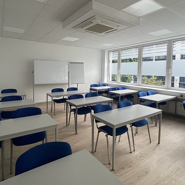 Heller Unterrichtsraum mit weißen Tischen, blauen Polsterstühlen, Holzboden. Im Hintergrund stehen ein Flipchart und ein Whiteboard.