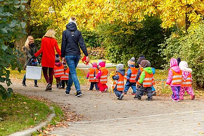 Drei Erzieherinnen machen mit ihrer Gruppe von Kita-Kindern einen herbstlichen Spaziergang im Park. Die Blätter der Bäume erstrahlen bereits in herbstlichen Farben. Die Kinder sind wetterfest und mit reflektierenden Sicherheitswesten angezogen und gehen den Erzieherinnen hinterher. 
