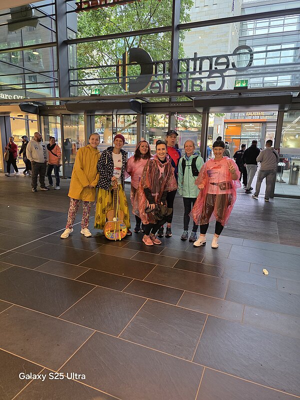 Eine Gruppe von acht Personen in Laufkleidung posiert im Eingangsbereich der Centrum Galerie. Einige tragen bunte Regenponchos, eine Gitarre steht in der Mitte.