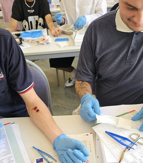 Lernen unter Rekordhitze: Praktische Übungen mit erfrischender Pausenversorgung Zwei Männer mit blauen Einmalhandschuhen üben die Wundversorgung. Auf dem Arm eines Teilnehmers ist eine künstliche Wunde angebracht, während medizinisches Material auf dem Tisch liegt.