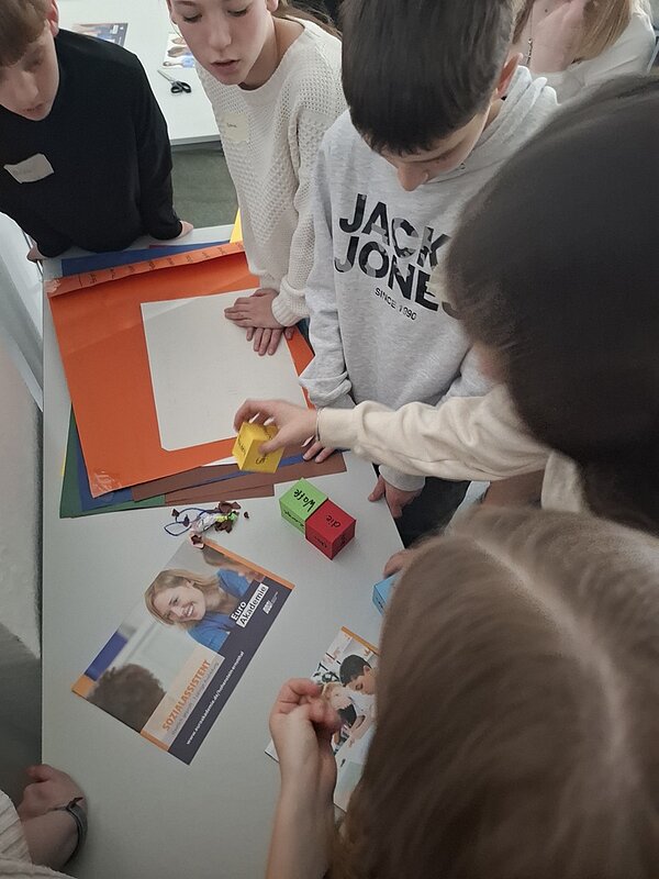 Schülerinnen und Schüler stehen um einen Tisch herum, auf dem Lernwürfel liegen, ein Euro Akademie Informationsflyer und Bastelpapier.