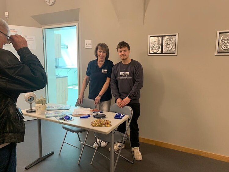 Interessierte Besucher*innen Eine Mitarbeiterin im T-Shirt mit Logo der Euro Akademie und ein Schüler stehen in einem Schulflur hinter einem Tisch, auf dem Broschüren und Give-Aways liegen. Ein Mann, der gerade seine Brille anhebt, ragt links ins Bild rein.