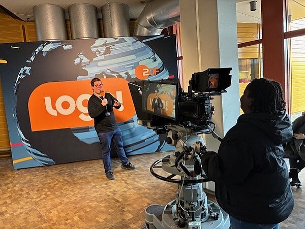 Ein junger Mann mit Mikrofon steht vor einer Wand mit der Aufschrift logo ZDF und wird dabei gefilmt.