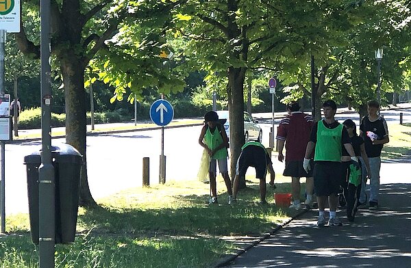 Eine Gruppe von Personen sammelt auf einem Gehweg neben einer Straße Müll auf. Einige tragen grüne Westen und halten Müllsäcke.