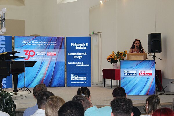 Euro Akademie Görlitz und Zittau: Feierliche Exmatrikulation Vor dem Altar in einer Kirche ist ein Podium aufgebaut und verschiedene mobile Wände mit dem Euro Akademie Logo. Am Podium steht eine Frau und hält eine Rede.
