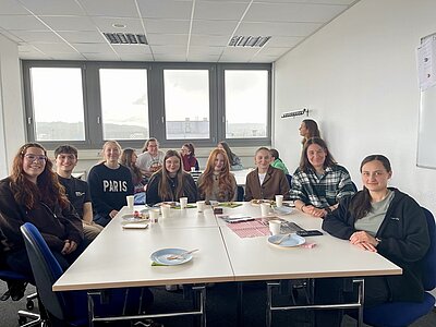 Die Gruppe der Alumni sitzt an einem Tisch und genießt eine Mahlzeit in einem hellen Raum der Euro Akademie Aschaffenburg. Alle sind gut gelaunt, lächeln und posieren für ein Gruppenfoto. 