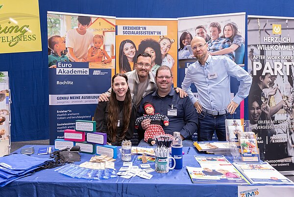 Am Messestand der Euro Akademie Rochlitz sitzen und stehen drei Männer und eine Frau; ein Mann hat eine Puppe auf dem Arm; vor ihnen liegen Info- und Werbematerialien auf einem Tisch mit blauer Tischdecke.