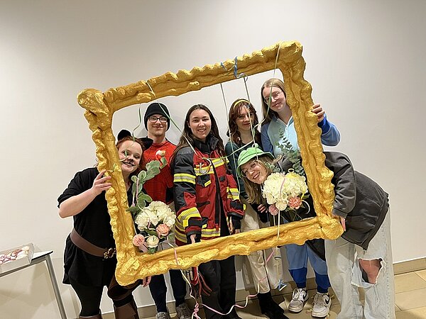 Sechs Studierende schauen lächelnd in ihren Faschingskostümen durch einen übergroßen selbst gestalteten Bilderrahmen und posieren für ein Foto.