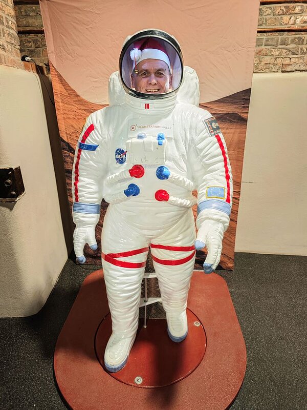 Eine Person steht lächelnd in einem Raum und trägt ein weißes Astronautenkostüm mit roten und blauen Akzenten. Das Kostüm hat ein Helmvisier und verschiedene Aufnäher, darunter ein NASA-Logo. Die Person trägt außerdem eine rote Weihnachtsmütze auf dem Kopf unter dem Helm. Im Hintergrund sind ein Wandbild mit einer Marslandschaft und ein weißer Kasten zu sehen.
