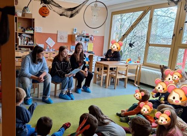 Drei Erwachsene sitzen auf kleinen Stühlen vor im Halbkreis auf dem Boden knieenden Kindern. Die Kinder haben bei der Bildbearbeitung ein stilistisches Mausgesicht auf die Gesichter gelegt bekommen, um ihre Identität zu schützen. Ein weitere Erwachsener, der weiter hinten im Raum sitzt hat ebenfalls ein "Mausgesicht", um anonym zu bleiben. Eine der erwachsenen Personen hält ein Buch in den Händen und scheint daraus vorzulesen. Die Einrichtung des Raumes lässt auf eine Kindertagesstätte schließen.