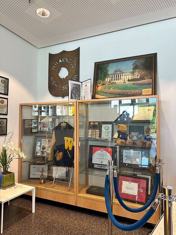 Ausstellungsvitrinen in einem Schulgebäude, gefüllt mit Trophäen, Urkunden, einem Kleidungsstück und anderen Erinnerungsstücken. Über den Vitrinen hängt ein Schild mit der Aufschrift 'H.H. Arnold High School' und ein gerahmtes Bild eines Gebäudes.