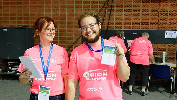 Zwei lächelnde Personen in pinken Volunteer-T-Shirts mit Namensschildern stehen in einer Halle; eine Person hält einen Ausweis in die Kamera.