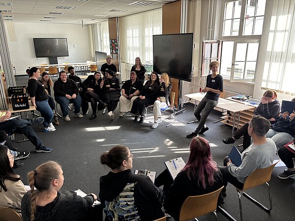 Pflegemesse Diabetes an der Euro Akademie In einem Klassenzimmer hat sich ein großer Stuhlkreis mit Auszubildenden versammelt. Die Schüler*innen blicken in Richtung der Dozentin, die vor einem geöffneten Fenster an einem Tisch lehnt und spricht. Im Klassenzimmer gibt es mehrere digitale Bildschirme