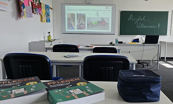 Im Unterrichtsraum stehen Tische und blau gepolsterte Stühle, auf einem Tisch liegen zwei Lehrbücher zum Thema Pflege, auf der Tafel steht "Herzlich willkommen".