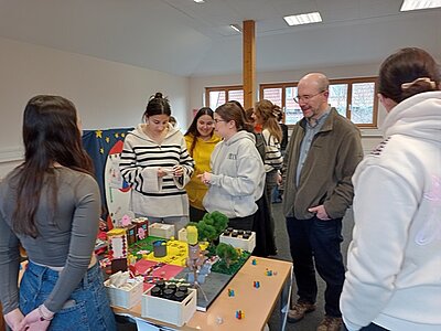 Lehrkraft und Auszubildende stehen vor einem Tisch einer Auszubildenden, die ihre Halbjahresarbeit in Form eines 3D-Brettspiels präsentiert.