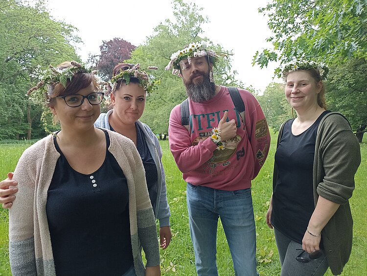 Blumenkränze basteln Drei junge Frauen und ein junger Mann grinsen in die Kamera. Sie tragen alle vier Blumenkränze auf ihren Köpfen. Der Herr in der Runde zeigt einen "Daumen hoch". Im Hintergrund sieht man eine Wiese und Bäume.