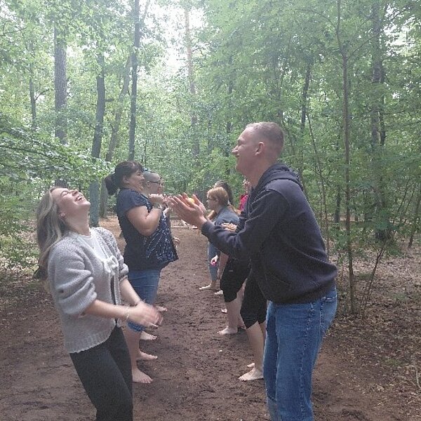 Eine Gruppe von Menschen steht in zwei Reihen sich gegenüber auf einem Waldweg und interagiert miteinander. Einige klatschen in die Hände, während andere lachen. Der Hintergrund zeigt dichte Bäume und grünes Laub.