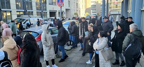 Euro Akademie und Euro-Schulen on Fire! Eine große Gruppe von Menschen steht vor einem Gebäude, das mit 'Euro Akademie' beschriftet ist. Die Personen tragen warme Kleidung und scheinen sich in einer Wartesituation zu befinden. Autos und Fahrräder sind im Hintergrund zu sehen, ebenso wie ein Gehweg und Straßenschilder.
