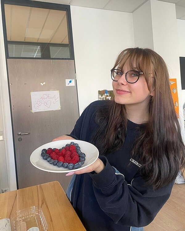 Weibliche Auszubildende mit langen braunen Haaren und Brille hält lächelnd ihren Teller für ein Foto hin, auf dem ein Herz aus Himbeeren und Blaubeeren liegt.